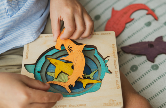 Child placing colorful wooden sea animal puzzle pieces (shark and fish) into a wooden board while sitting on a bed.